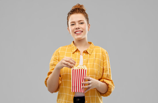 fast food and people concept - smiling red haired teenage girl in checkered shirt eating popcorn from striped bucket over grey background