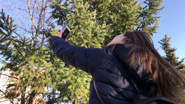 Slowmo of young girl taking selfies with a smartphone using front camera in a city park. Back medium shot.