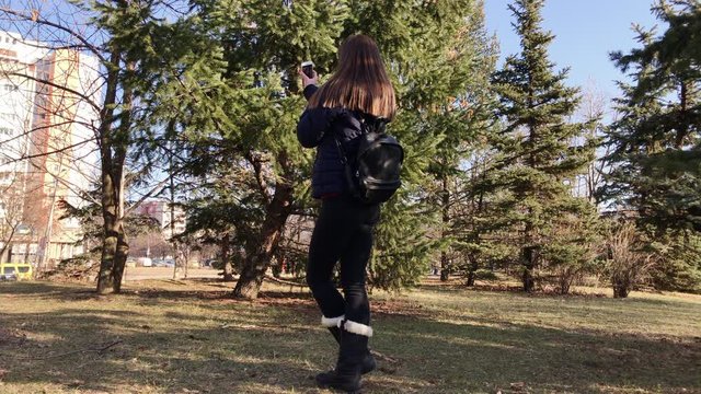 Young girl taking selfies with a smartphone using front camera in a city park. Wide back shot.