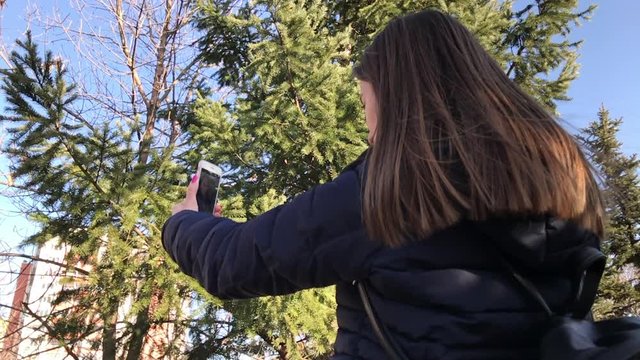 Slowmo of young girl taking selfies with a smartphone using front camera in a city park. Back panning shot.