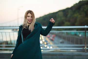 Positive young woman wearing green coat posing at the bridge with lights. Space for text