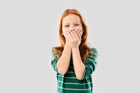 childhood, embarrassment and emotions concept - confused red haired girl in green striped shirt covering her mouth by hands over grey background
