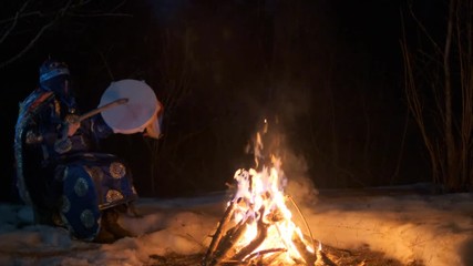 The shaman beats his drum sitting near the fire.