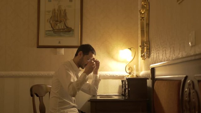 Man in white shirt finish writing taking off the glasses and thinking over a wooden desk.