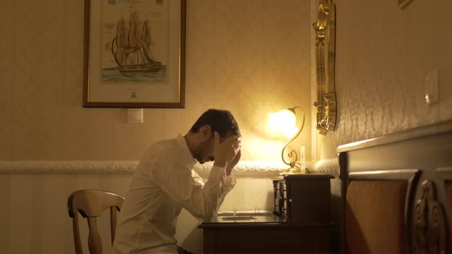 Man in white shirt sadly thinks about writing something. Then he puts on his glasses and writes on a wooden desk.