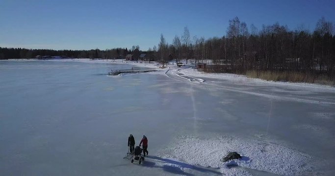Aerial, drone shot of young mother with baby stroller and senior woman with kick sled walking on icy lake on a sunny winter day