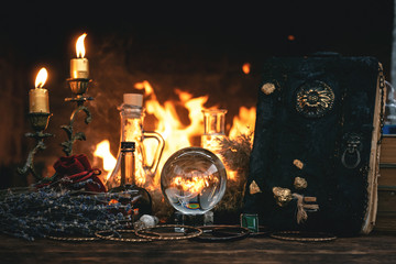 Crystal ball, magic book, magic potion and other wizard accessories on a table on a burning fire background.