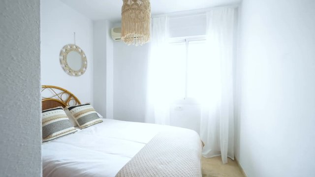 Camera enteres in a comfortable hotel bedroom with rattan headboard with natural fabric cushion, macrame mirror, fiber nightstand and a big window with view.Holiday destination apartment.