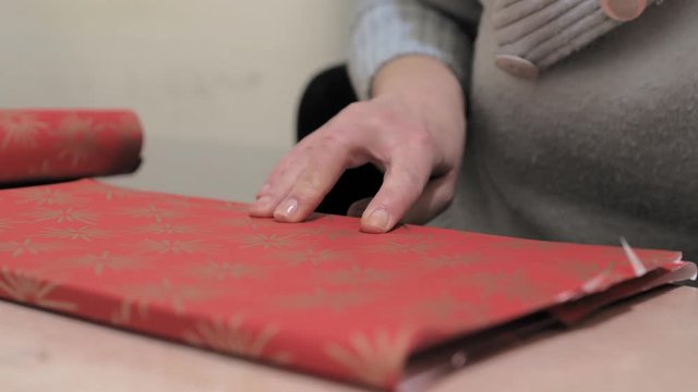 Young woman tapes down an edge of red wrapping paper on a gift; wrapping Christmas presents.