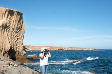 Elderly caucasian female traveling into Tenerife island looking at the horizon over water with binocular. Exploring and enjoying the nature. Windmill on background