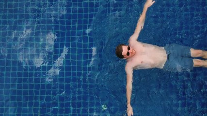 White man wearing blue shorts and sunglasses swimming calmly in a blue pool