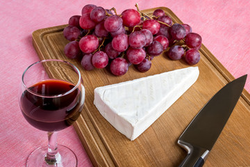 Glass of red wine and a triangular piece of Brie cheese, red sweet grapes, chef knife on a brown wooden cutting board. Soft cheese covered with edible white mold.