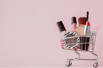Creative concept with shopping trolley with makeup on a pink background. Perfume, sponge, brush,...