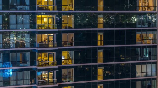 Glowing apartment windows at night in glass skyscraper timelapse. Warm light and motion inside. Lights reflected from surface. Zoom out