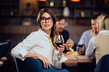 Portrait of gorgeous brunette looking away and holding glass with red wine while sitting at restaurat. In background friends eating diner.
