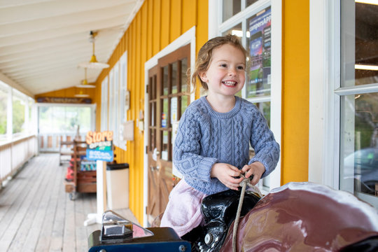 A young girl riding on a toy horse in front of a small town store.