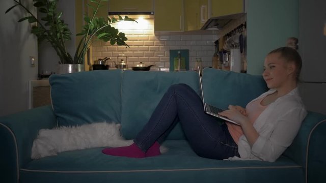 A young woman uses the laptop sitting on the couch in the evening with a cat. 4K