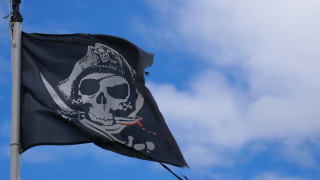 Black ragged pirate flag with bloody knife between skull teeth and crossed swords waving on the pole against blue sky and white clouds