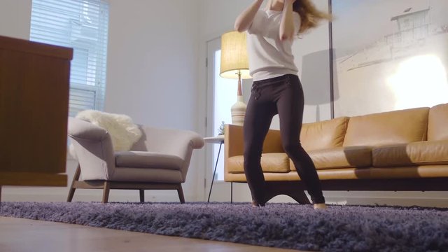 Slow motion ground level view of a young woman having fun dancing in her lounge room, 23.98 fps.