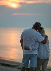 A middle-aged couple hugging each other and observing a sunset sea, we see them from the back