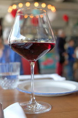 Low angle close-up shot of clear wine glass with traditional round goblet shape filled with dark red wine on wood tabletop with blurry restaurant background scene