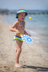 A boy in striped pants and panama hat plays ping-pong with a striped racket, standing near the sea on sand