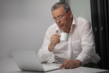 A middle-aged grey-haired man in a white shirt, sitting at a table with a cup in his hand, looking in a notebook