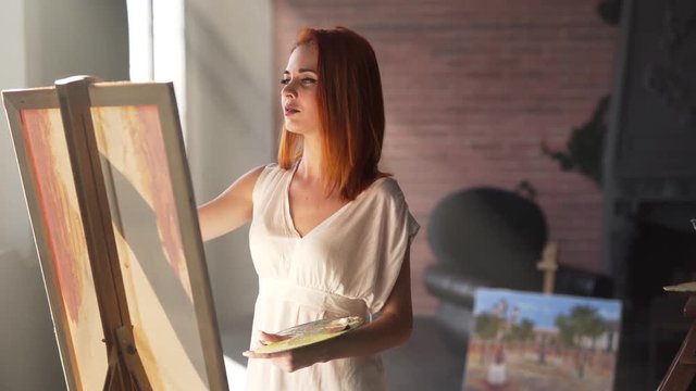 Seductive young red-haired girl is standing in her workroom in only white transparent shirt and painting. There are a lot of sunrays in a room.