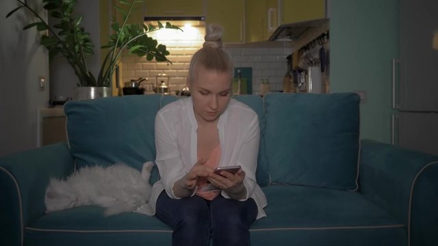 A young woman uses the phone sitting on the couch in the evening with a cat. The girl read the good news on the phone. 4K