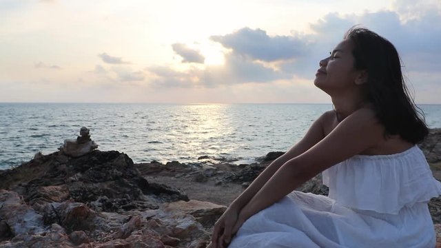 A young thai lady is sitting at a rocky shore during sunset, cloes her eyes, smiles, and shakes her hair while they float on the wind then stops and opens her eyes. She enjoys the moment and is happy.