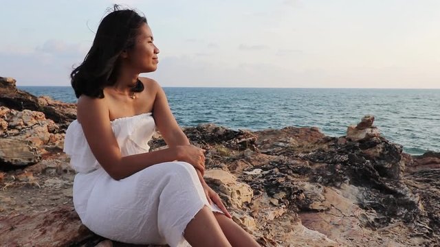 A young thai lady in a white dress is sitting at the rocks near the ocean and waiting for a sunset while taking a deep breath and closing her eyes  enjoying her moment, Thailand.