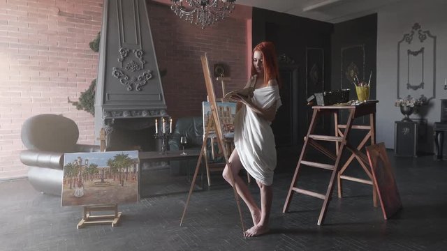 Seductive young red-haired girl is standing in her workroom in a white shirt and painting. The workroom is made in a loft style.