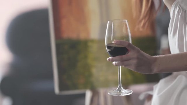 Focused on a glass of wine. Seductive young red-haired girl is sitting on a chair in front of a picture she is painted holding a glass of red wine and correcting something with a brush and paints.