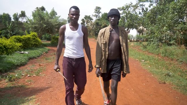 A front facing tracking shot of two young African gangsters with weapons walking down a rural dirt road in Africa.