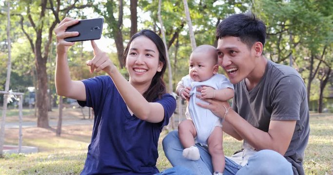 Mother and Father using smartphone to selfie with baby at park. Asian family having quality time together.