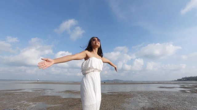 A young thai woman in a white dress is making a circle around herself with her arms extended, smiling and closing her eyes showing how happy she is on a beautiful beach.