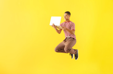 Portrait of jumping African-American teenage boy with laptop on color background