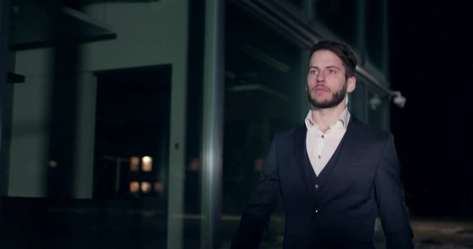 Businesman walks by night in a city through urban street next to a glass building in a suit camera is sliding next to him.