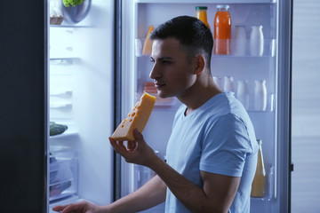 Handsome man eating tasty cheese near refrigerator at night