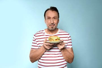 Portrait of handsome mature man with tasty burger on color background