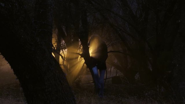 A woman looks paranoid as she is walking slowly between trees in the woods on a cold night, 23.98 fps.