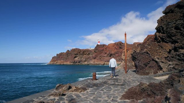 View of a tourist walking at the dock, then stops and takes a photo of the volcanic cliffs and the famous lighthouse of Teno in the Isle of Tenerife. Slow motion edit