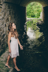 Alone beautiful girl with curly natural hair in white dress in ancient flooded tunnel with stony walls. Lonely princess in old castle. Dreamer young girl in long tunnel with water. Moss in old tunnel.