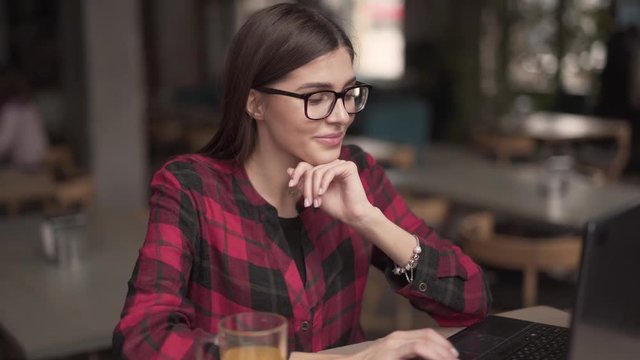 An attractive brunette sits in the cafe and types on her laptop. She smiles and is in the good mood. She places her hand gracefully under her chin.  Waiter passes by on the background that is blurred