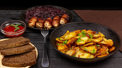 Roasted potato in the pot, pieces of rye black bread on a wooden serving plate, homemade sausages in bacon and stewed cabbage on a black rustic background