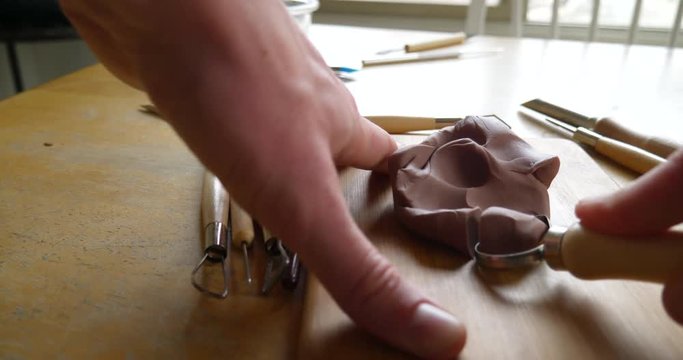 An artist carving and cutting brown modeling clay with a metal tool before sculpting and shaping the clay with his fingers SLIDE RIGHT.