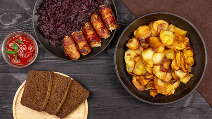 Roasted potato in the pot, pieces of rye black bread on a wooden serving plate, homemade sausages in bacon and stewed cabbage on a black rustic background