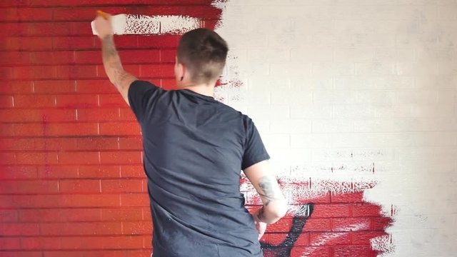 Tattooed artist painting over a red mural on a brick wall with white paint