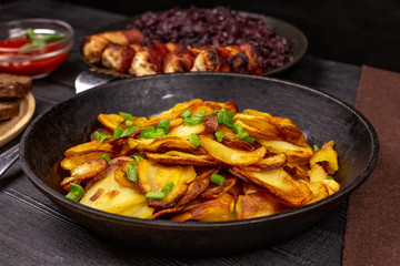 Fried potato chips with greens in a pot, in front of homemade sausages and stewed cabbage, on a black rustic background