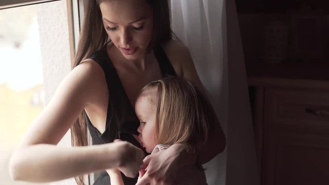 Young Caucasian Mother With Long Black Hair and Black Dress Breast Feeding Her Baby Girl Daughter By The Window and Looks Outside